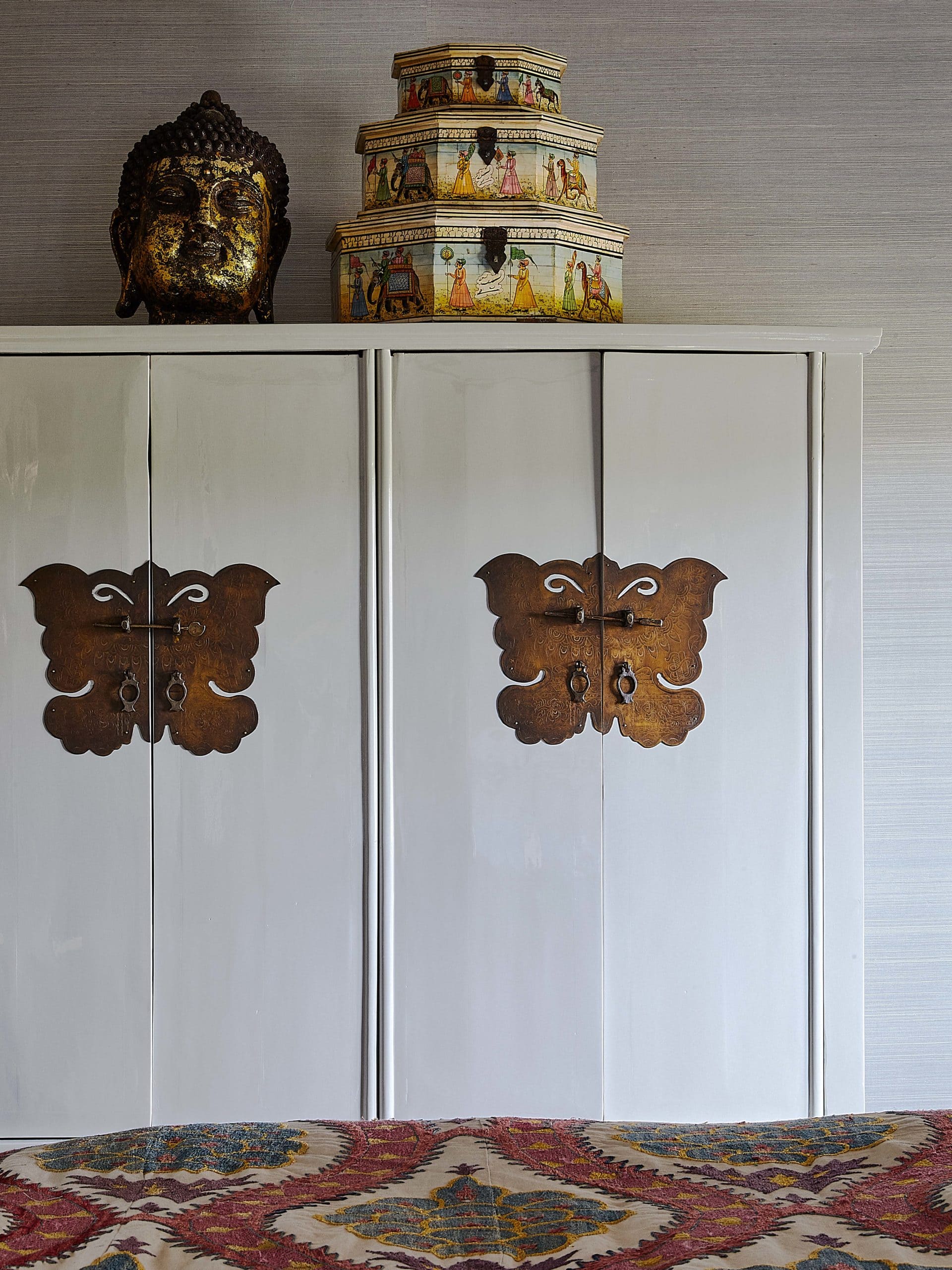 White asian cabinet in bedroom with painted bone hat boxes atop. Brass Buddha head decor.